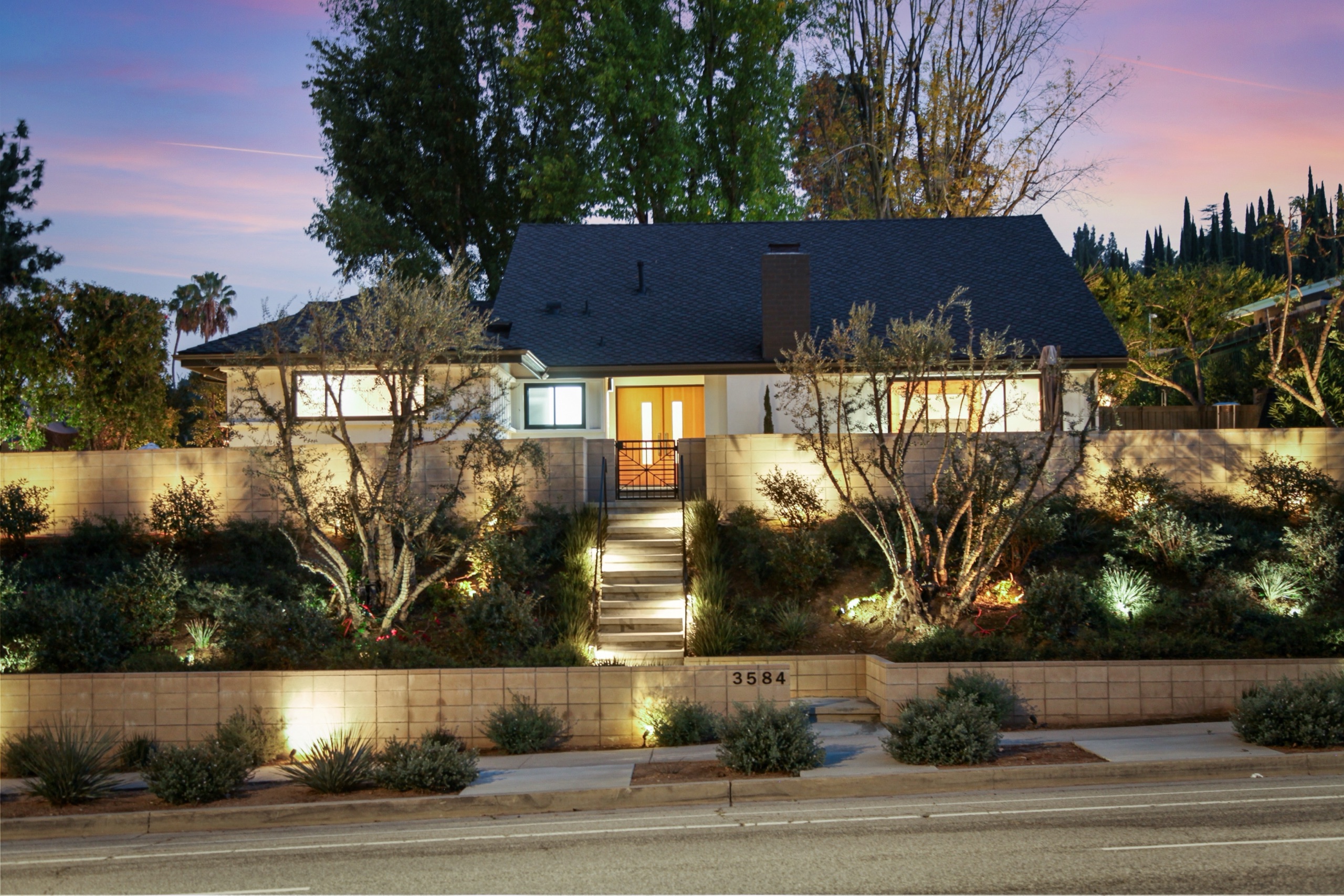 3584 Sepulveda Blvd, Sherman Oaks, at twilight. A 1962 mid-century modern residence with a dark gabled roof, warm glowing entry doors, and terraced landscaping against a rose and lavender sunset sky.